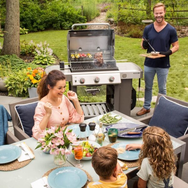 Familia al aire libre disfrutando de una comida mientras un hombre cocina en una barbacoa Weber Genesis II E-310 GBS