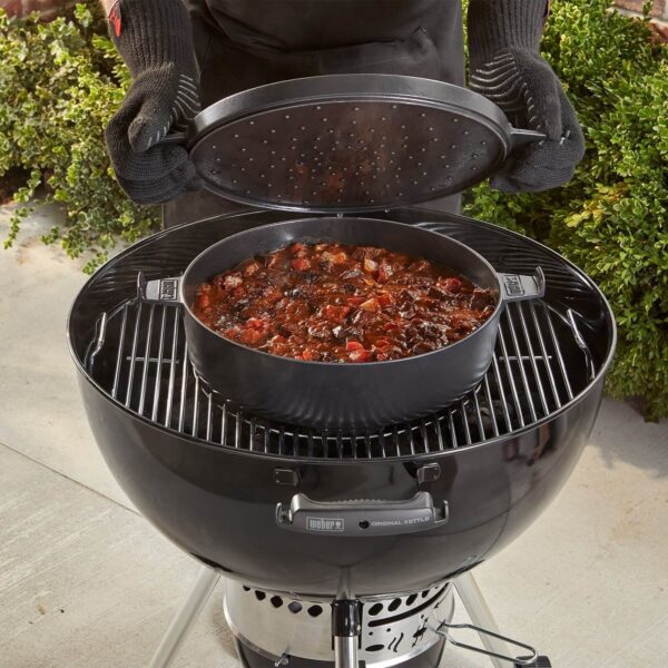 Persona levantando la tapa perforada de una olla de Hierro Colado Weber en una barbacoa, mostrando un guiso cocinándose.