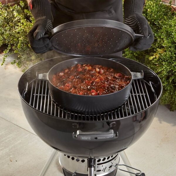 Persona levantando la tapa de la olla de Hierro Colado Weber en una barbacoa, revelando un guiso cocinándose.