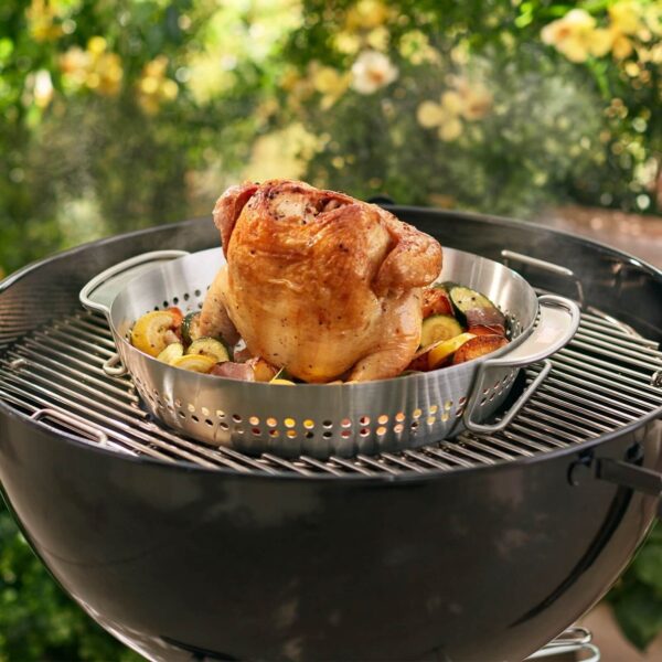 Pollo asado en posición vertical sobre un soporte Weber de acero inoxidable, acompañado de mazorcas de maíz y otras verduras, cocinándose en una barbacoa Weber.