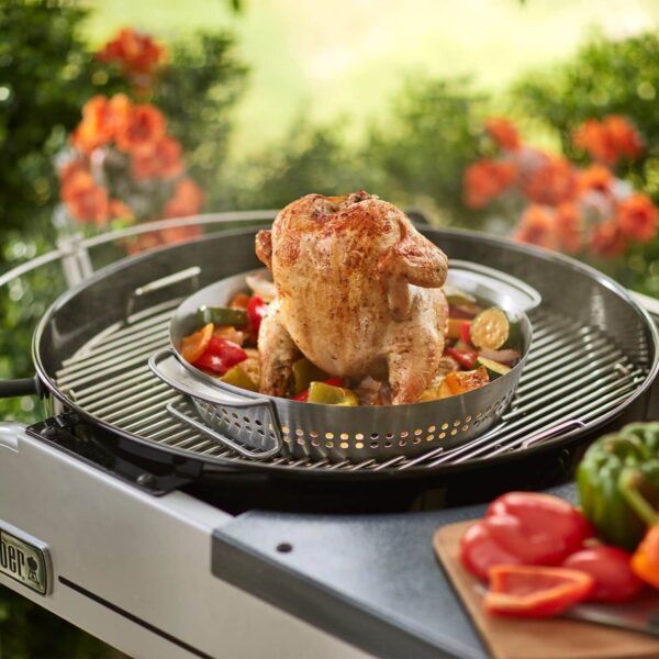 Pollo asado en posición vertical sobre un soporte Weber, rodeado de verduras frescas, cocinándose al aire libre en una barbacoa.