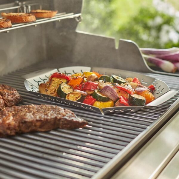 Verduras asadas coloridas en cesta Weber junto a jugosos filetes en una barbacoa de gas.