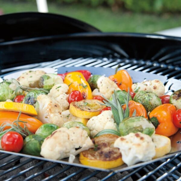 Variedad de verduras asadas en cesta de acero inoxidable Weber sobre una parrilla al aire libre.