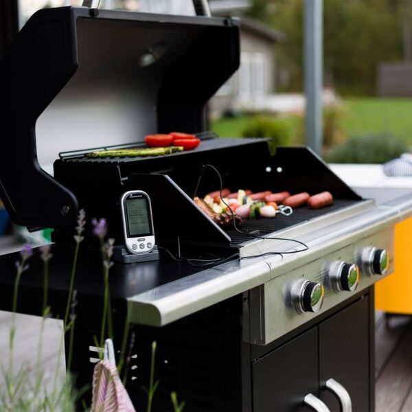 Parrilla de gas en uso con verduras y salchichas cocinándose y un termómetro digital en primer plano.