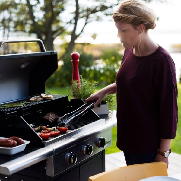 Mujer cocinando en una parrilla de gas al aire libre en un jardín al atardecer.