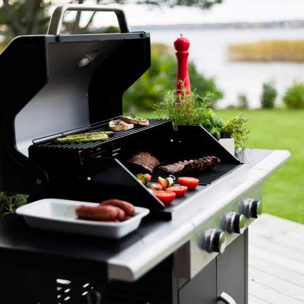 Parrilla de gas cocinando carnes y verduras en un jardín con vista al lago al atardecer.