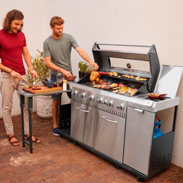 Dos amigos disfrutando de cocinar al aire libre en una barbacoa de gas Rexon 6.1, mostrando la mesa lateral y la amplia superficie de cocción en uso.