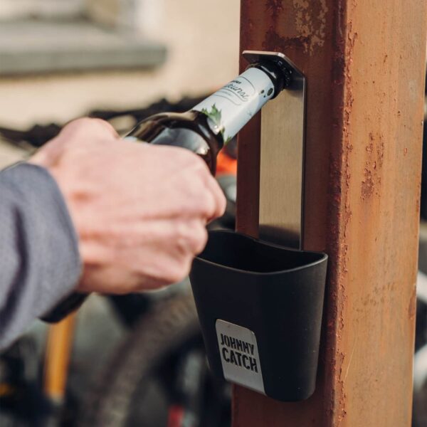 Mano de una persona utilizando el abrebotellas Johnny Catch montado en una columna oxidada para abrir una botella de cerveza.