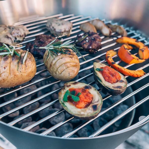 Diversos vegetales y carnes asándose en la parrilla de una barbacoa CONE de Höfats, destacando las posibilidades de una cocina al aire libre diversa.