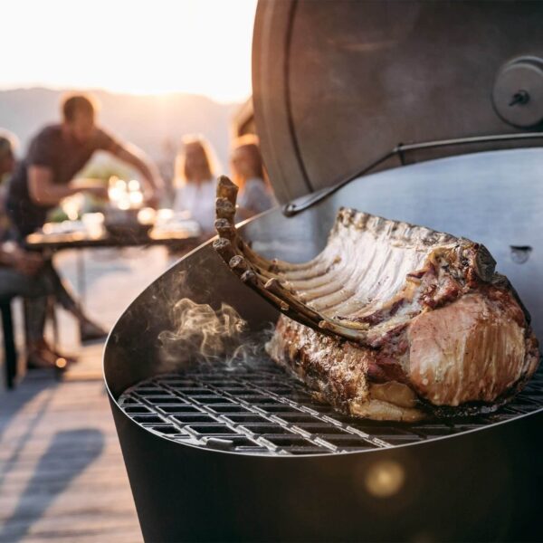 Un jugoso costillar cocinándose en una barbacoa CONE de Höfats al atardecer con amigos reunidos al fondo.