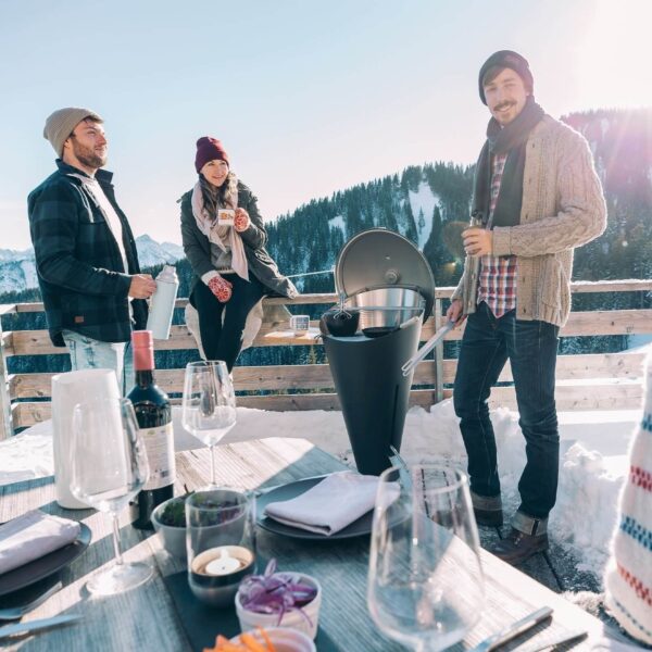 Grupo de amigos disfrutando de bebidas calientes alrededor de una barbacoa CONE de Höfats en un entorno montañoso nevado.