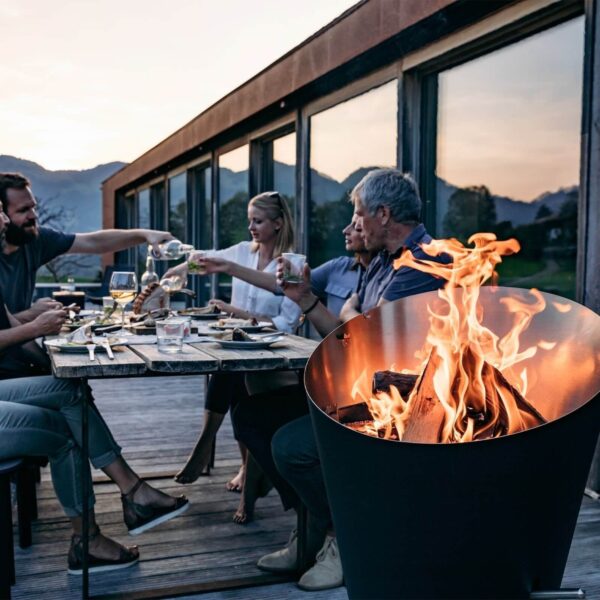 Amigos brindando alrededor de una cálida cesta de fuego Höfats en una terraza durante el atardecer.