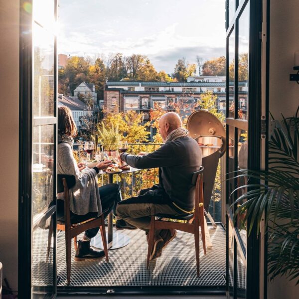 Pareja disfrutando de un almuerzo en un balcón soleado con vistas a un paisaje urbano otoñal y una barbacoa CONE de Höfats.