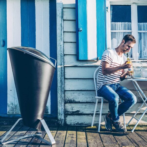 Hombre sonriente disfrutando de una bebida refrescante al lado de su barbacoa CONE de Höfats en una terraza soleada.