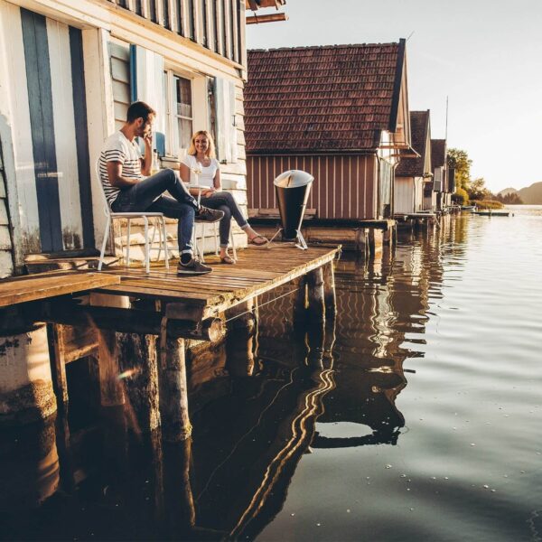 Pareja relajada conversando en un muelle de madera con la barbacoa CONE de Höfats a la orilla del lago al atardecer.