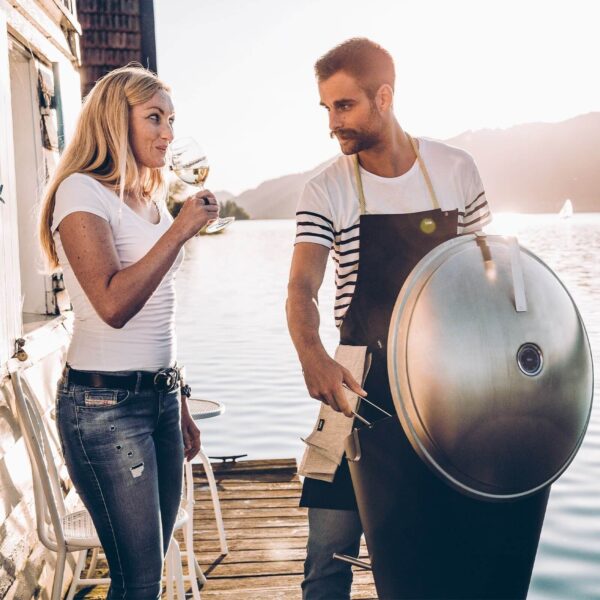 Pareja relajándose junto al lago con una copa de vino y una barbacoa de carbón CONE de Höfats al atardecer.