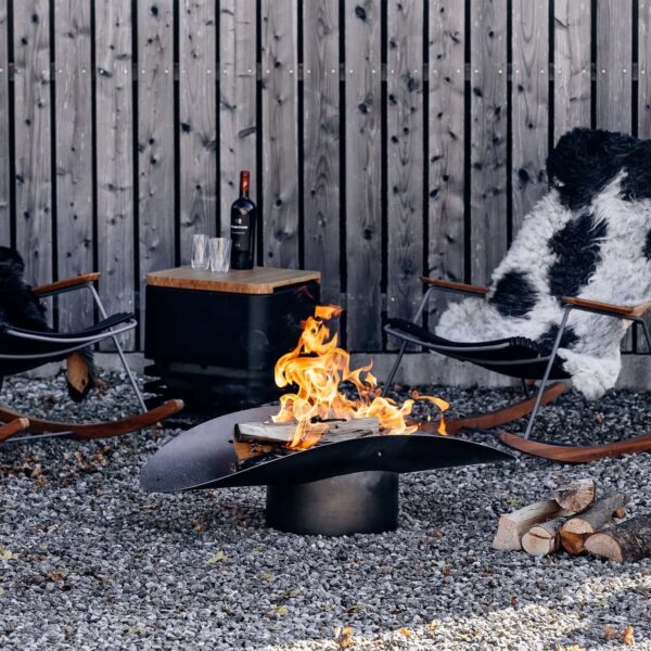 Vivo fuego ardiendo en el Brasero Ellipse de Höfats en una terraza con sillas y mesa de madera, creando un ambiente acogedor.