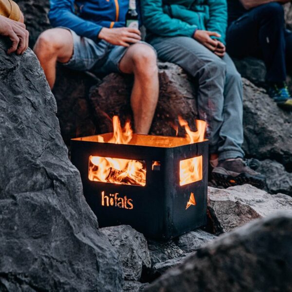 Grupo de amigos disfrutando de un cálido fuego con el BEER BOX Black de Höfats en una playa rocosa al atardecer.