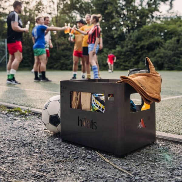 BEER BOX Black de Höfats utilizado como almacenamiento de objetos personales en un campo de fútbol con jugadores al fondo.