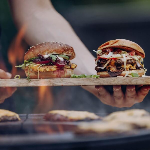 Manos sosteniendo una tabla de madera con hamburguesas gourmet frente a una parrilla de fuego al aire libre.