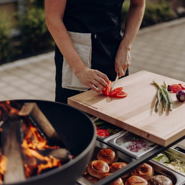 Chef preparando ingredientes frescos en una encimera de madera junto a una parrilla de fuego al aire libre.