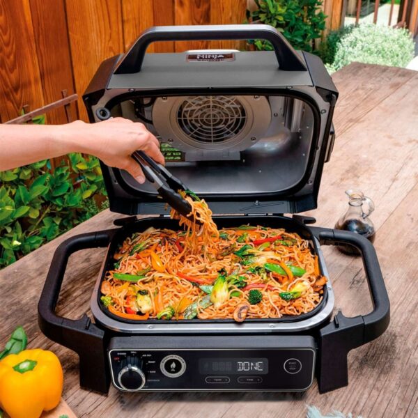 Una persona cocinando un delicioso plato de noodles vegetales en la Barbacoa Eléctrica Ahumador Woodfire en un entorno al aire libre.