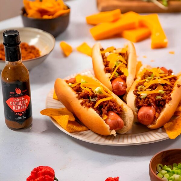 Placa con tres chili dogs cubiertos con carne picada, queso y cebolla, acompañados de nachos. A la izquierda, una botella de salsa Carolina Reaper de Doctor Salsas, en tonos negros y rojos.