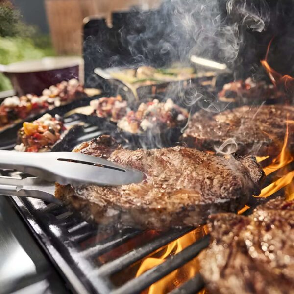 Primer plano de filetes y verduras cocinándose en la barbacoa Weber Genesis E-415 con pinzas de cocina