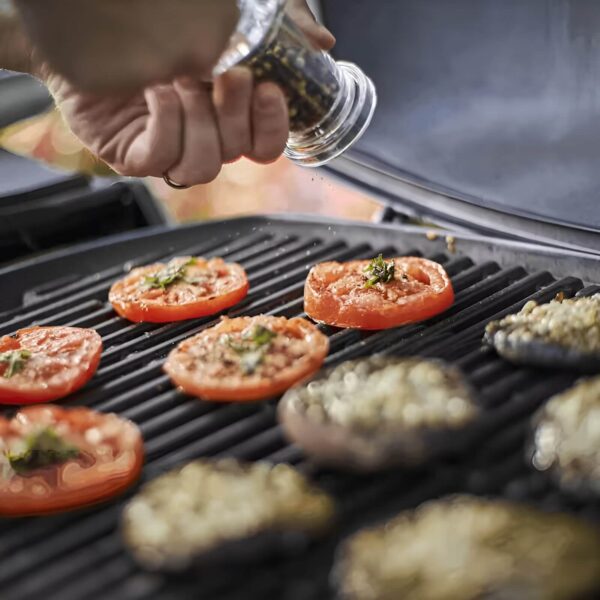 Mano sazonando rodajas de tomate sobre la parrilla de la barbacoa Weber Q 2200 durante la cocción.