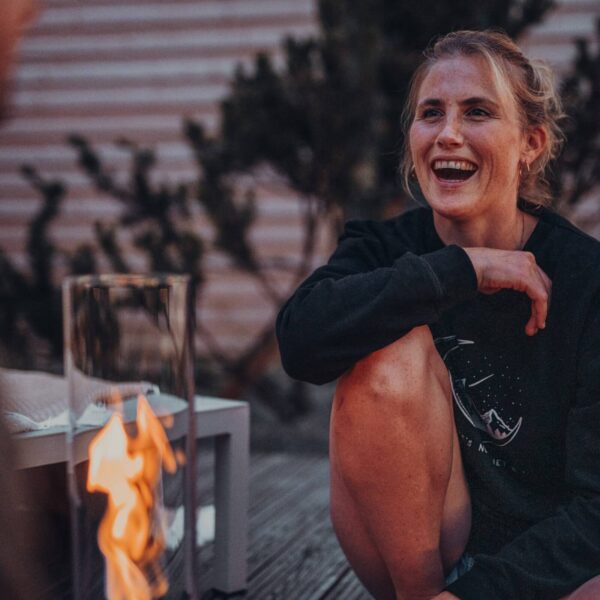 Una mujer sonriente se calienta junto a un fuego de mesa Spin 120 en un ambiente al aire libre al atardecer.