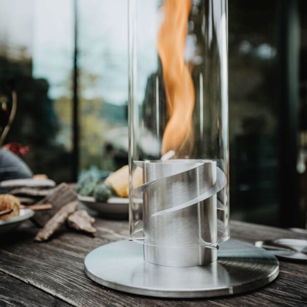 Una lámpara de fuego de mesa Spin 120 en acabado plateado sobre una mesa de madera rústica al aire libre, con naturaleza desenfocada de fondo.