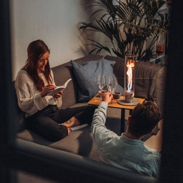 Una mujer leyendo un libro y un hombre sirviendo una bebida junto a una lámpara de fuego de mesa Spin 90, proporcionando un ambiente íntimo.