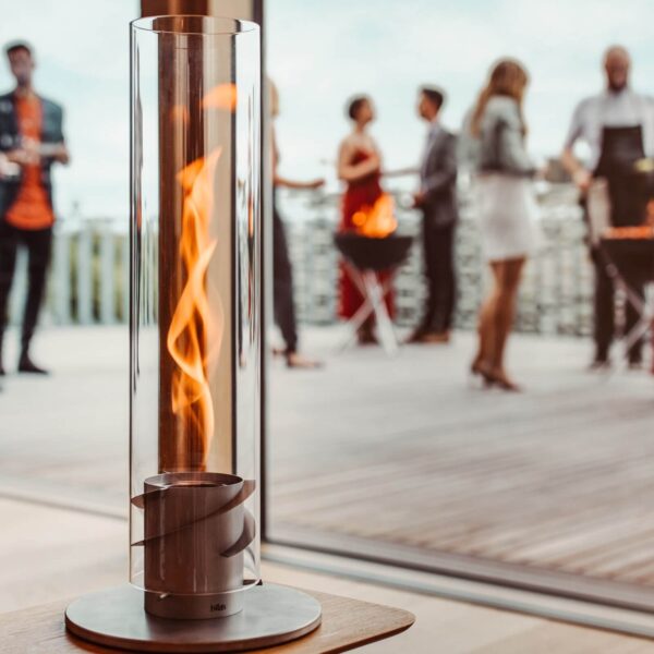 Una lámpara de fuego de mesa Spin 120 de diseño elegante en una terraza de madera con personas socializando al fondo.