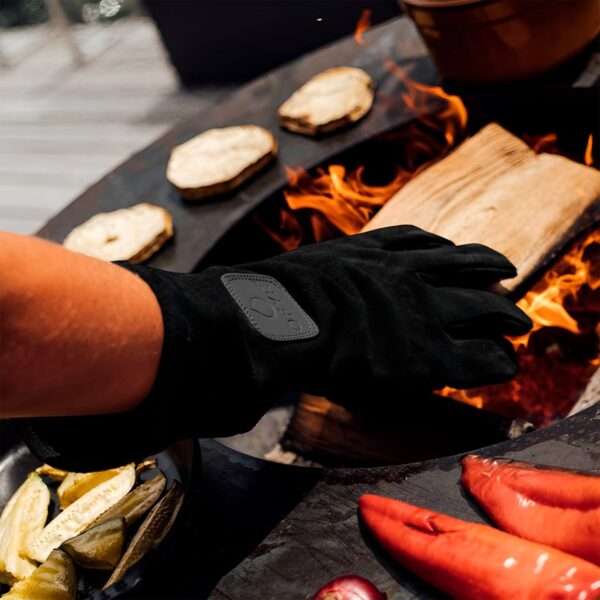 Persona usando guantes de gamuza negros para manipular alimentos en una parrilla encendida, mostrando la protección contra el calor al cocinar al aire libre.