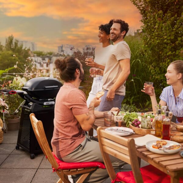 Personas disfrutando de una comida al aire libre junto a una barbacoa Weber Q 2800N+ con carro negro en una terraza al atardecer.