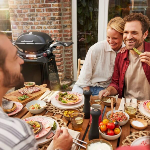 Grupo de personas disfrutando de una comida al aire libre junto a una barbacoa Weber Q 2800N+ en un patio acogedor.