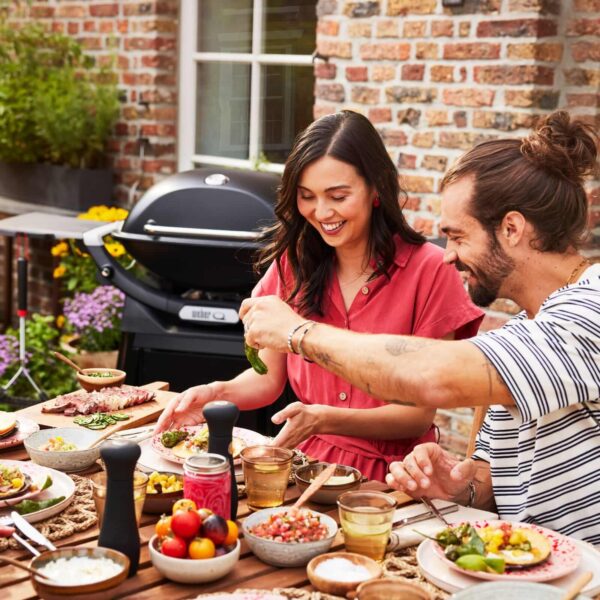 Pareja disfrutando de una comida al aire libre junto a una barbacoa Weber Q 2800N+ en un patio lleno de plantas.