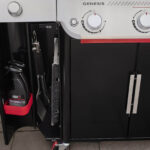 Compartimiento de almacenamiento lateral en la barbacoa Weber Genesis EP-335W con utensilios y productos de limpieza organizados.
