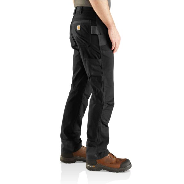 Hombre de perfil usando pantalones técnicos Carhartt negros con refuerzos y botas de trabajo.