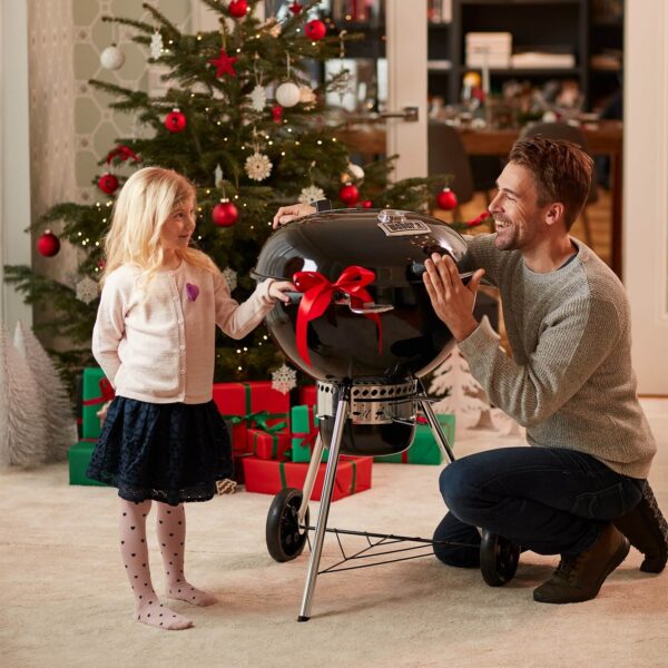 Niña y hombre sonrientes junto a una barbacoa Weber Master-Touch Premium decorada con un lazo rojo, frente a un árbol de Navidad.