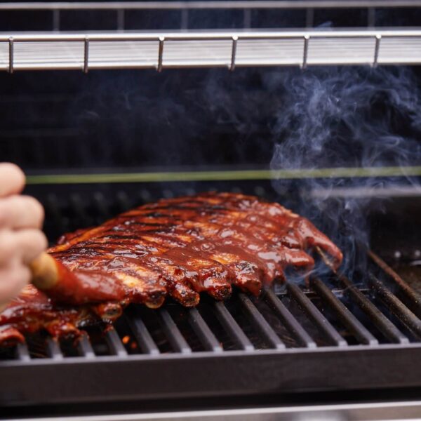 Mano aplicando salsa BBQ sobre costillas asándose en la parrilla de la barbacoa BeefEater Discovery 1200E, con humo visible.