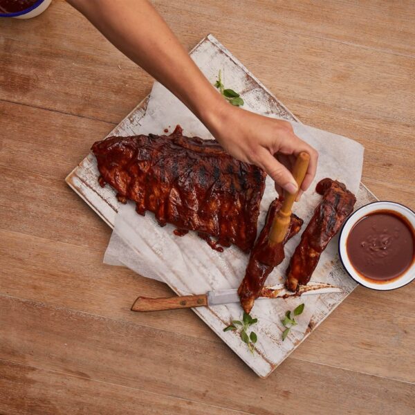 Mano aplicando salsa BBQ sobre costillas a la parrilla, servidas sobre una tabla de madera con un cuenco de salsa.