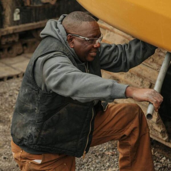 Hombre trabajando al aire libre usando un chaleco Carhartt negro, con capucha gris y pantalones marrones