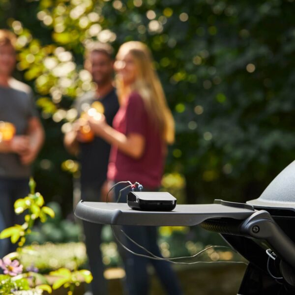 Weber Connect Smart Grilling Hub en una parrilla al aire libre con personas disfrutando en el fondo.