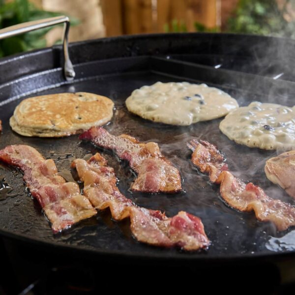 Tiras de beicon dorándose junto a panqueques con arándanos en una plancha de hierro fundido sobre una barbacoa Weber Kettle.