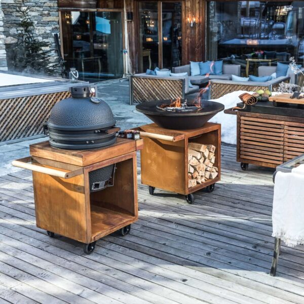 Mesa Kamado OFYR en terraza de montaña, con barbacoa Kamado negra y leña almacenada.