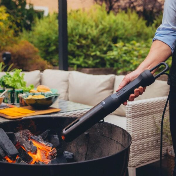 Persona usando el Looft Air Lighter 2 para encender carbón en una parrilla al aire libre, con un fondo de jardín y mesa preparada.