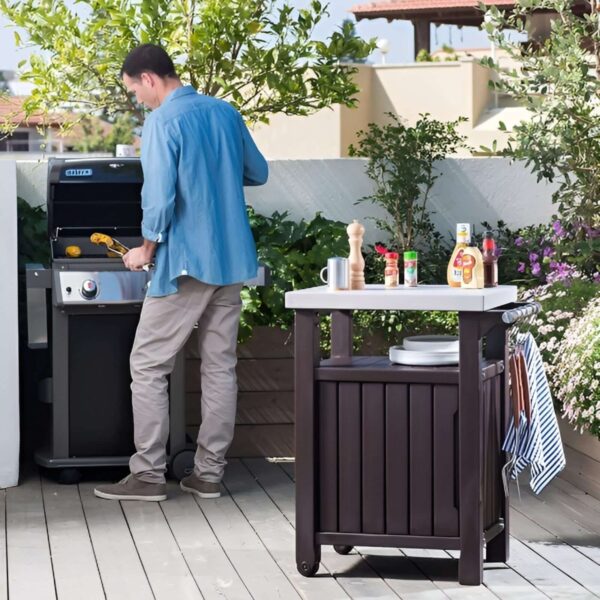 Hombre cocinando en una parrilla junto a un mueble auxiliar Keter, que sostiene platos, condimentos y utensilios, en una terraza al aire libre.