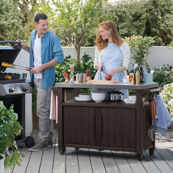 Pareja cocinando al aire libre con un mueble auxiliar Keter grande, utilizado para organizar utensilios, platos y condimentos durante la preparación de alimentos.