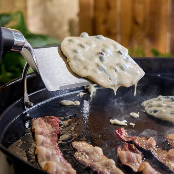 Espátula de acero inoxidable levantando una tortita con arándanos en una plancha caliente mientras se cocinan tiras de tocino.